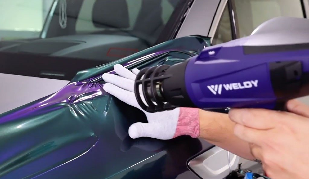  person uses a heat gun to smooth out a colorful vinyl wrap on a car hood, ensuring a tight fit and removing wrinkle