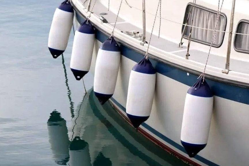 Boat fenders hanging from the side of a sailboat, reflecting in the water.